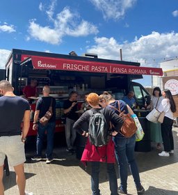 Foodtruck mit frischer Pasta und Pinsa Romana zieht hungrige Besucher an – sonniger Tag, rote Markise und entspannte Atmosphäre auf einem Streetfood-Markt oder Festival.