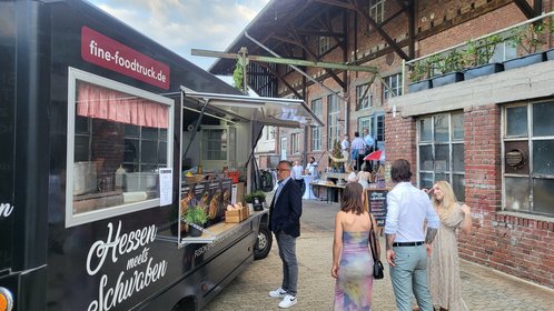 Elegantes Streetfood-Event mit Fine-Foodtruck „Hessen meets Schwaben“ vor Backsteinhalle – Gäste in festlicher Kleidung genießen regionale Spezialitäten in stilvollem Ambiente.