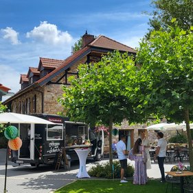 Foodtruck vor historischem Gebäude bei Sommerwetter, Gäste stehen an Stehtisch auf gepflegtem Rasen unter Bäumen.
