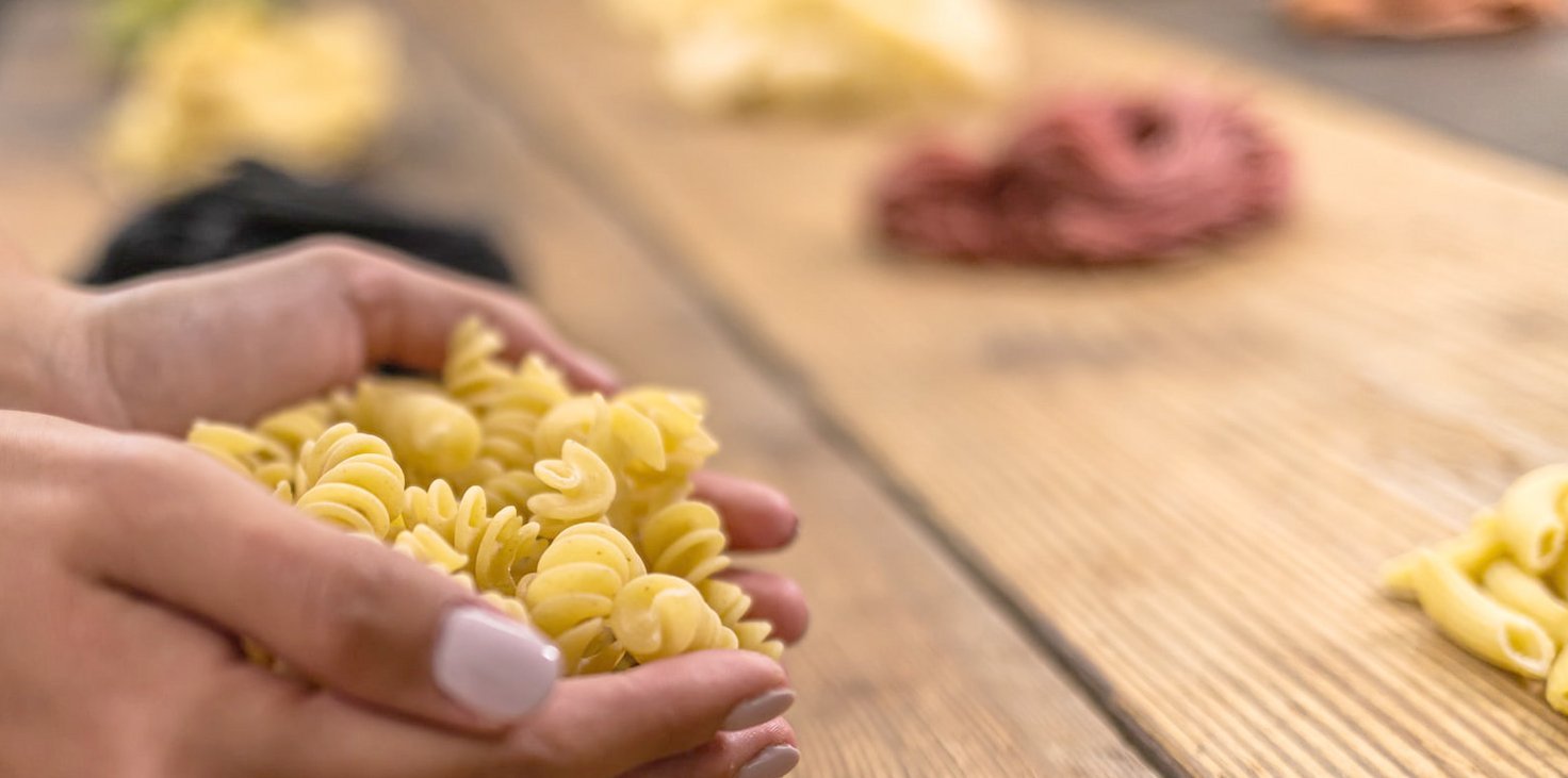 Nahaufnahme von Händen, die rohe Fusilli-Nudeln halten, mit weiteren Pasta-Sorten unscharf im Hintergrund auf einem Holztisch.