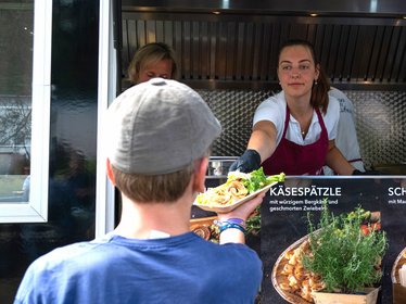 Frau im Foodtruck reicht einem Jungen eine Portion Käsespätzle mit Röstzwiebeln und Salat bei Essensausgabe.