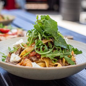Frische Tagliatelle mit Rindfleisch, Tomatensoße, Rucola und Ricotta in Keramikschale, im Hintergrund Pizza mit Burrata.
