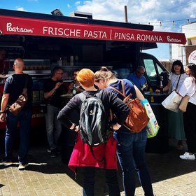Menschen stehen vor einem roten Foodtruck mit Aufschrift „Frische Pasta | Pinsa Romana“ bei sonnigem Wetter.