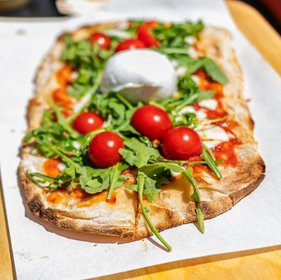 Knusprige Fladenpizza mit Rucola, Kirschtomaten und cremigem Burrata auf Backpapier serviert – mediterrane Streetfood-Spezialität bei Sonnenschein.