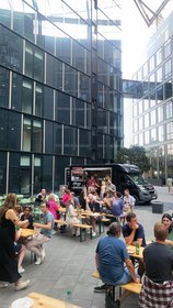 Menschen genießen Streetfood an Bierbänken vor einem schwarzen Foodtruck in einem modernen Innenhof mit Glasfassaden.