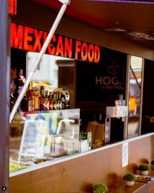 Mexikanischer Foodtruck mit rotem „Mexican Food“-Leuchtschild, Getränken und Gewürzen in der Auslage vor urbaner Kulisse.