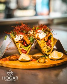 Zwei kreativ garnierte Tacos mit Fleisch, Gemüse und Sour Cream auf Holzbrett, serviert von Hogar Streetfood mit Limettenstücken.