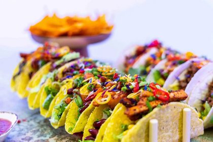 Bunte Reihe vegetarischer Tacos mit Bohnen, Paprika, Mais, Avocado und Chilis auf Tisch – im Hintergrund Schale mit Tortilla-Chips, ideal für Streetfood-Events oder Partys.