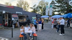 Besucher essen bei sonnigem Wetter an Tischen vor einem schwarzen Foodtruck mit italienischem Streetfood auf einem Platz mit Sonnenschirmen.