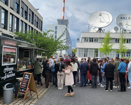 Menschen stehen bei einer Firmenveranstaltung vor einem Foodtruck mit dem Schriftzug "Hessen Schmücken", im Hintergrund Bürogebäude und Satellitenschüsseln.