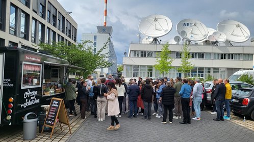 Lange Menschenschlange vor dem Foodtruck „Hessen meets Schwaben“ auf einem Firmengelände mit ARD-Satellitenschüsseln – beliebtes Streetfood-Event bei bewölktem Wetter.