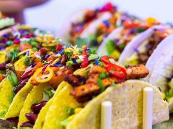 Bunte Tacos mit Gemüse, Bohnen, Grilltofu und frischen Chilis, kunstvoll angerichtet in Taco-Halterungen auf hellem Hintergrund.