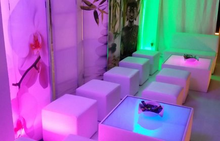 Moderne Lounge mit weißen Sitzwürfeln, farbiger LED-Beleuchtung in Pink und Grün, asiatischer Deko und Orchideen-Motiven an den Wänden.