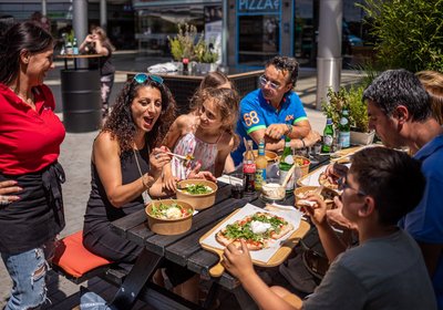Familie genießt italienisches Streetfood mit Pasta und Pizza an einem sonnigen Tisch im Freien vor einem modernen Restaurant.
