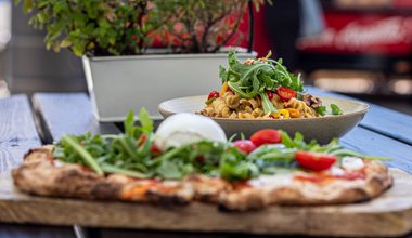 Frische Pizza mit Rucola, Tomaten und Burrata sowie ein Teller Pasta mit Gemüse auf Holztisch im Freien serviert.