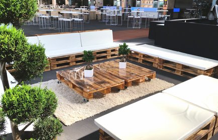 Outdoor Lounge mit weißen Polstern auf Palettenmöbeln, Holz-Palettentisch, Teppich und grünen Pflanzen in moderner Eventlocation.