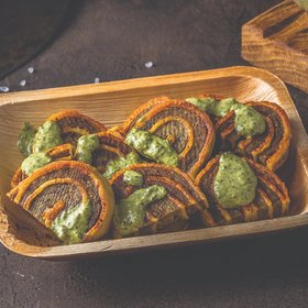 Vegetarische Linsenröllchen mit grüner Kräutersoße auf Holztablett, daneben frische Kräuter und ein Glas Dip auf dunklem Hintergrund.