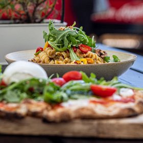 Italienische Pasta mit Rucola und Kirschtomaten im Fokus, davor unscharf eine Pizza mit Burrata, serviert auf Holzbrett im Freien.