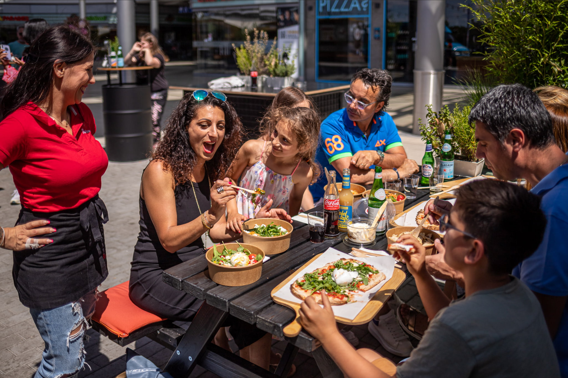 Fröhliche Gruppe genießt frische Pinsa, Salate und Softdrinks an einem sonnigen Tag im Außenbereich eines Restaurants – mediterranes Streetfood-Erlebnis mit Familie und Freunden.
